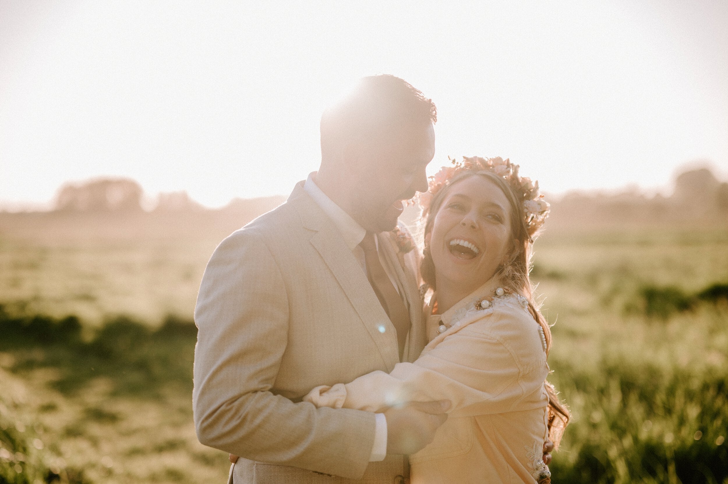 A couple embraces and laughs together in a sunlit field, wearing light-colored clothing. The woman, adorned with a floral crown in her hair, radiates joy. Captured by a talented Kent wedding photographer, this moment is pure magic.
