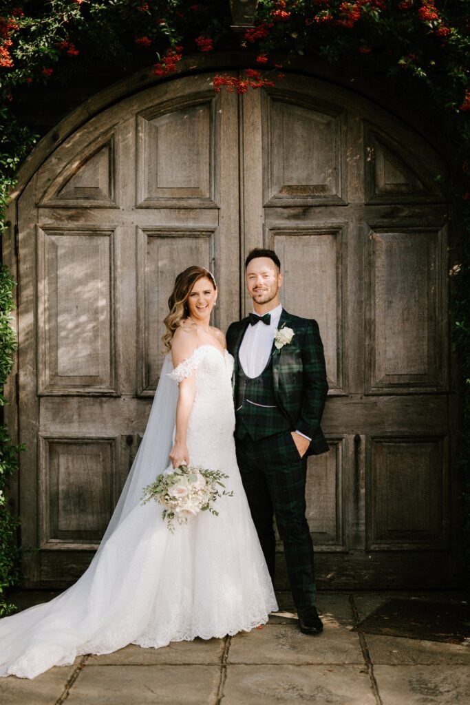 A bride in a white dress and groom in a dark green suit stand together at a charming Kent wedding venue, framed by a large wooden door decorated with greenery and red flowers.