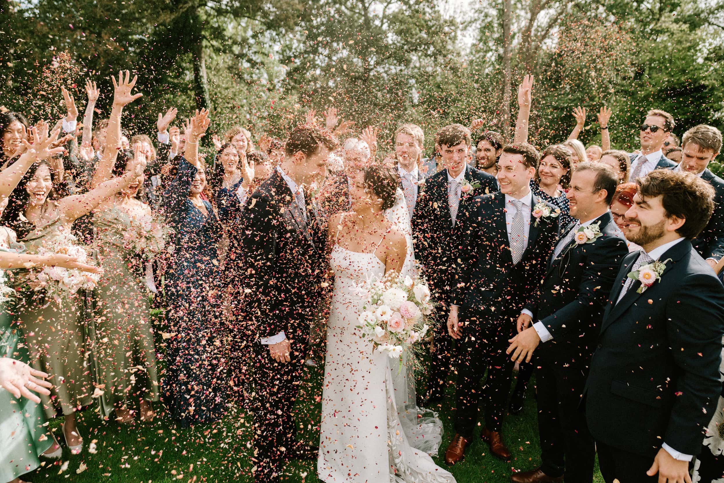 A newlywed couple stands surrounded by guests throwing confetti in a picturesque garden setting. The couple is smiling and holding hands, with trees in the background, captured beautifully by a skilled wedding photographer from Kent.