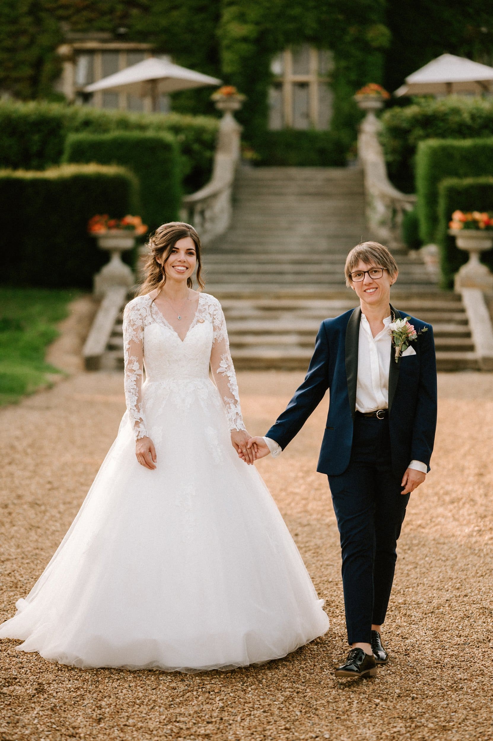 Two people, one in a wedding dress and the other in a suit, walk hand in hand outside a building with stairs and greenery, perfectly captured by their Kent wedding photographer.