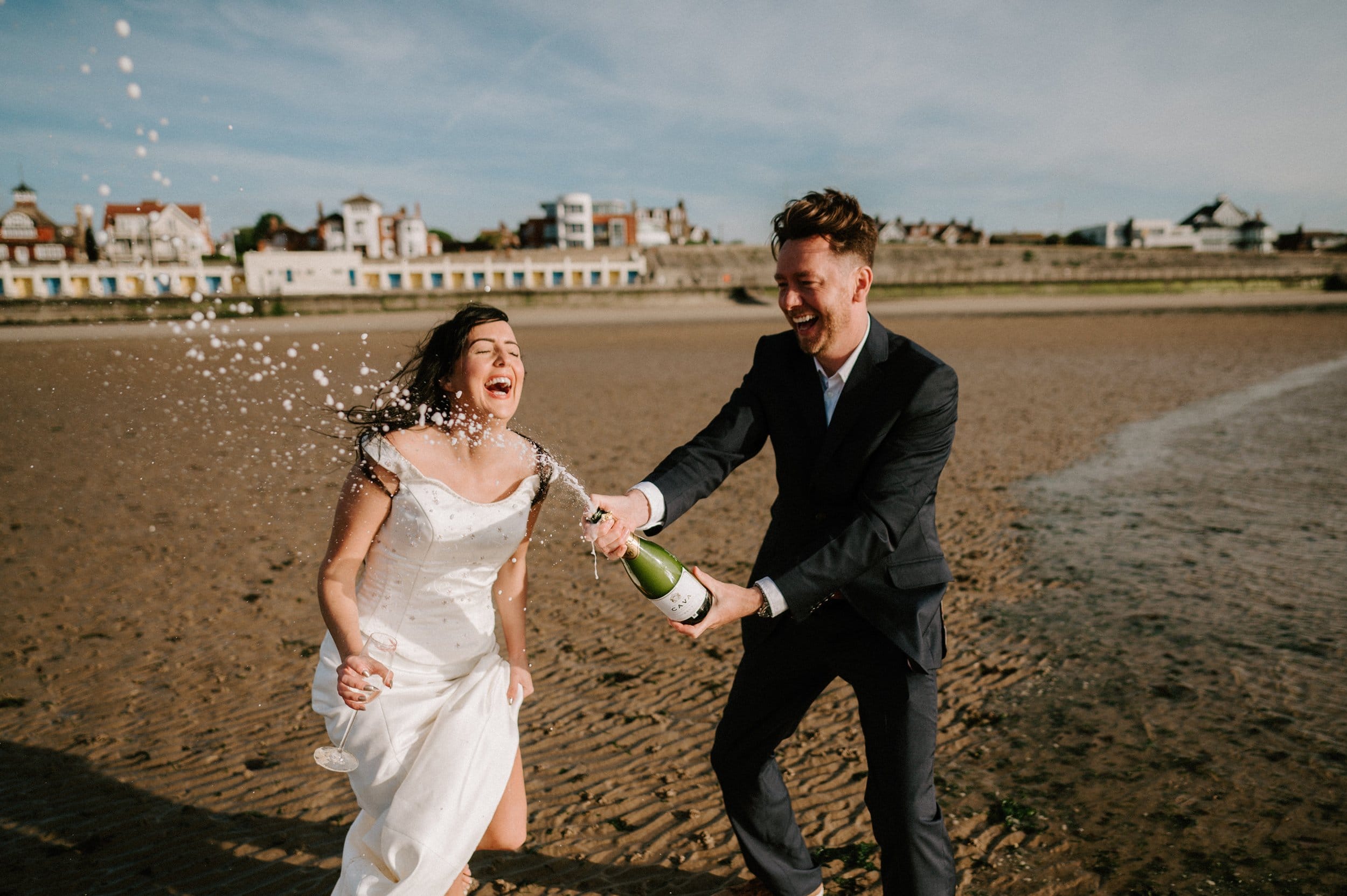 A woman in a white dress and a man in a suit laugh as he sprays champagne on a sandy beach. Several buildings are visible in the background along the shoreline, perfectly captured by their Kent wedding photographer.