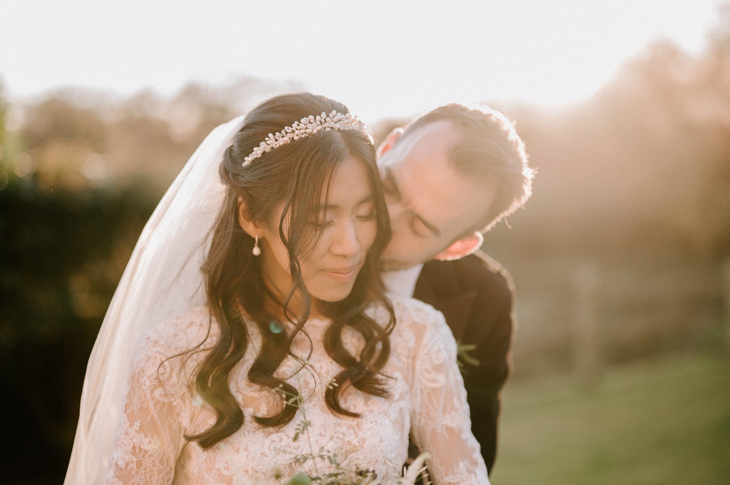 A newlywed couple shares an intimate moment outdoors at sunset. The bride, wearing a lace gown and tiara, is captured perfectly by their Kent wedding photographer, while the groom, dressed in a suit, embraces her from behind.