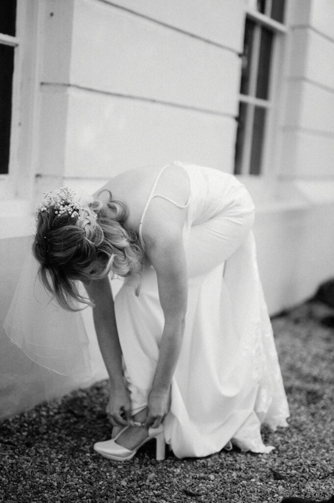 A woman in a wedding dress bends down to adjust her shoe near a building with large windows, perfectly capturing a candid moment by the skilled Kent wedding photographer.