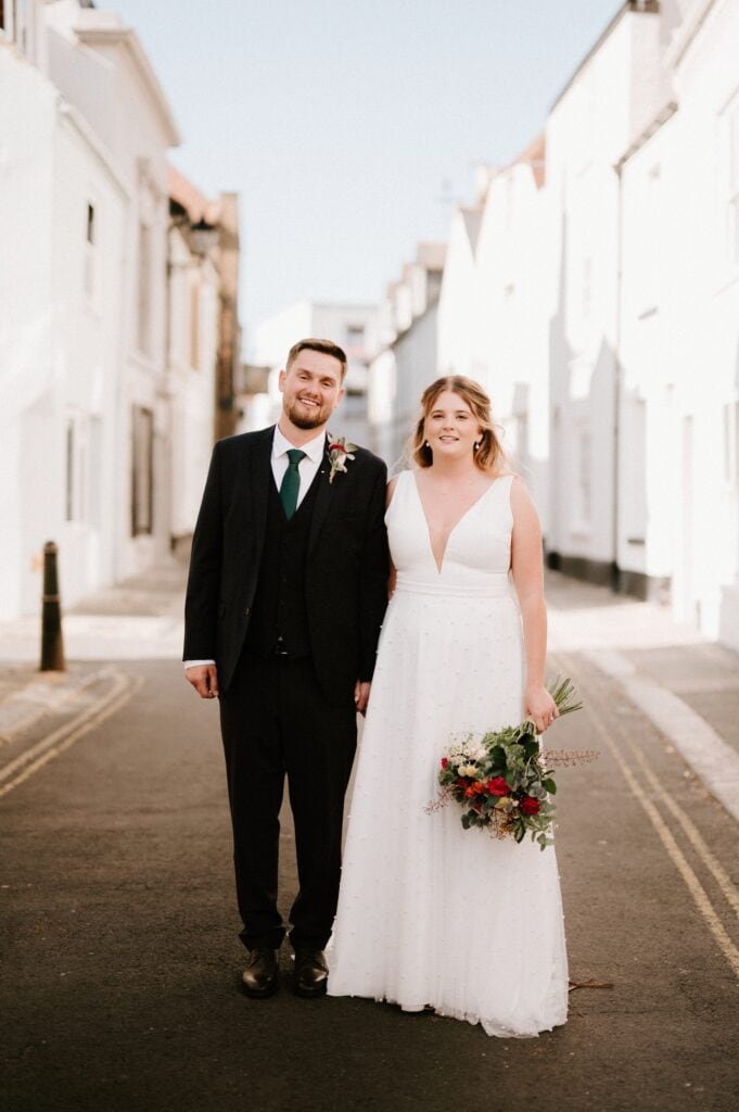 A man in a black suit and a woman in a white bridal gown stand side by side on a street between white buildings, with the woman holding a bouquet of flowers outside their dream Kent wedding venue.