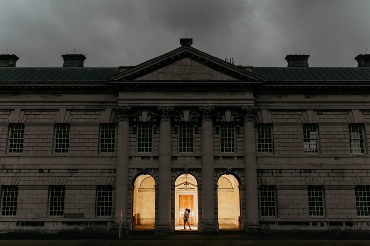 A couple stands illuminated under an archway of a large, grand building with columns at dusk, against a backdrop of a cloudy sky.