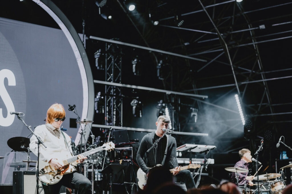 A band performs on an outdoor stage at night in Kent, showcasing two guitarists and a drummer, with various lighting and sound equipment in the background. A music photographer from London captures every electrifying moment.