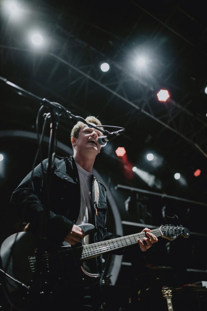 A musician in a dark jacket plays an electric guitar and sings into a microphone on a dimly lit stage in London. Bright lights shine from above as the moment is captured by a skilled music photographer from Kent.