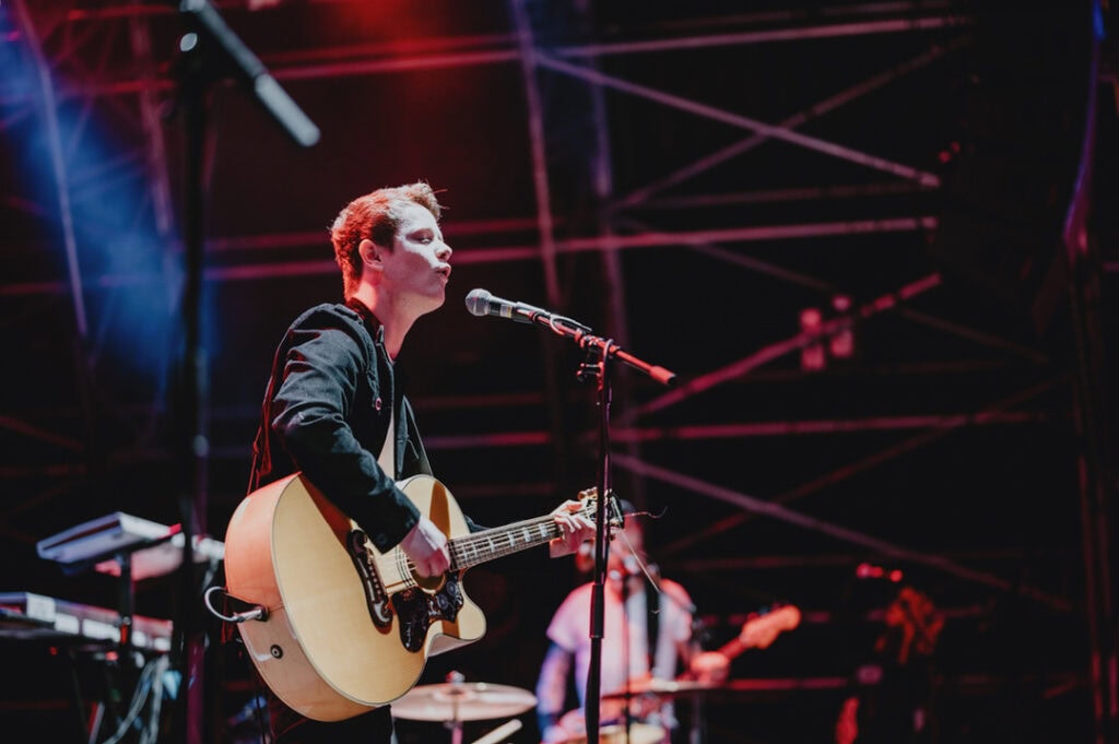 A person stands on a stage in London, playing an acoustic guitar and singing into a microphone. Scaffolding and stage lights are visible in the background, while a music photographer captures the moment.