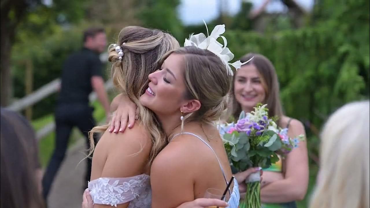Two women embrace at an outdoor event, their expressions joyful. One woman holds a bouquet, while several other people can be seen in the background.
