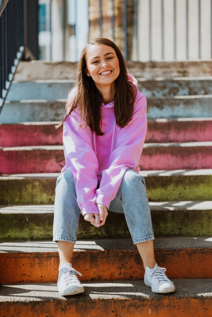 A person in a pink hoodie and jeans sits on outdoor steps, smiling at the camera.