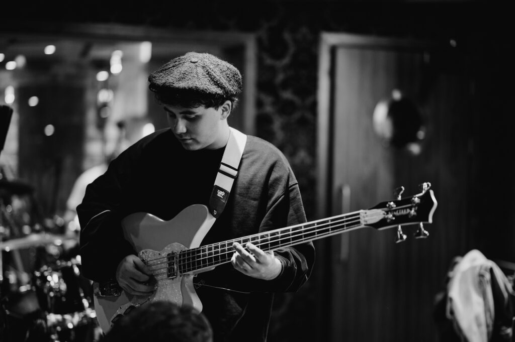 Black and white image of a person wearing a beret, intently playing an electric bass guitar in an indoor setting, captured by a renowned music photographer from London.