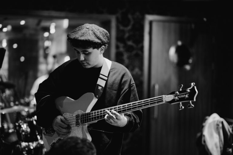 Black and white image of a person wearing a beret, intently playing an electric bass guitar in an indoor setting, captured by a renowned music photographer from London.