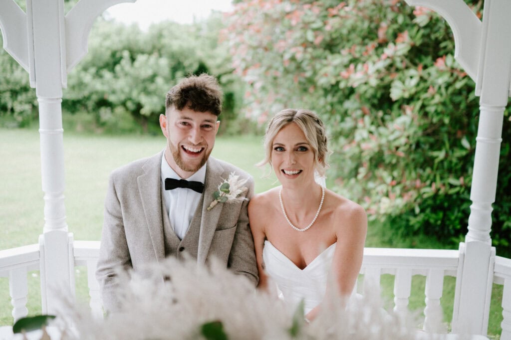 A smiling couple on their wedding day sits together under a white gazebo at an outdoor wedding venue in Kent. The groom wears a light gray suit with a black bow tie, and the bride dazzles in a white wedding dress with a pearl necklace. Greenery and flowers create a picturesque background. Image by Pearce Wedding Photography.