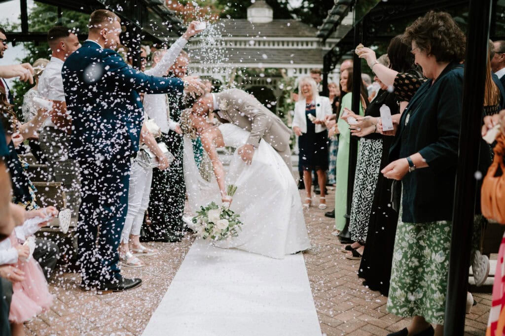 A newlywed couple shares a kiss on a white aisle at The Old Kent Barn as confetti is thrown by guests surrounding them. The groom, in a grey suit, leans the bride, in a white dress, backwards while she holds a bouquet of flowers. The scene is festive with smiles and celebratory confetti at their wedding. Image by Pearce Wedding Photography.