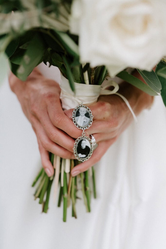 Close-up of a person's hands holding a bouquet of white roses. Two black and white photo lockets are attached to the bouquet.