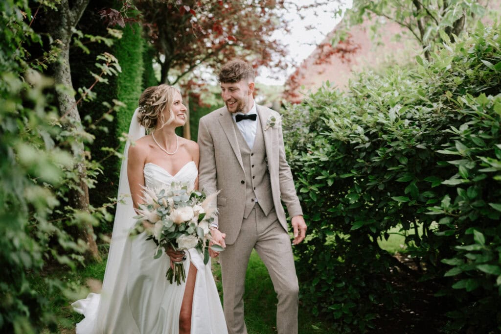 A bride and groom walk hand in hand through a garden. The bride holds a bouquet and wears a white dress, while the groom is in a beige suit.