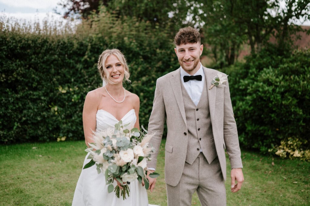 A bride and groom stand together outdoors, holding hands. The bride is wearing a white dress and holding a bouquet; the groom is in a beige suit with a black bowtie. Greenery is visible in the background.