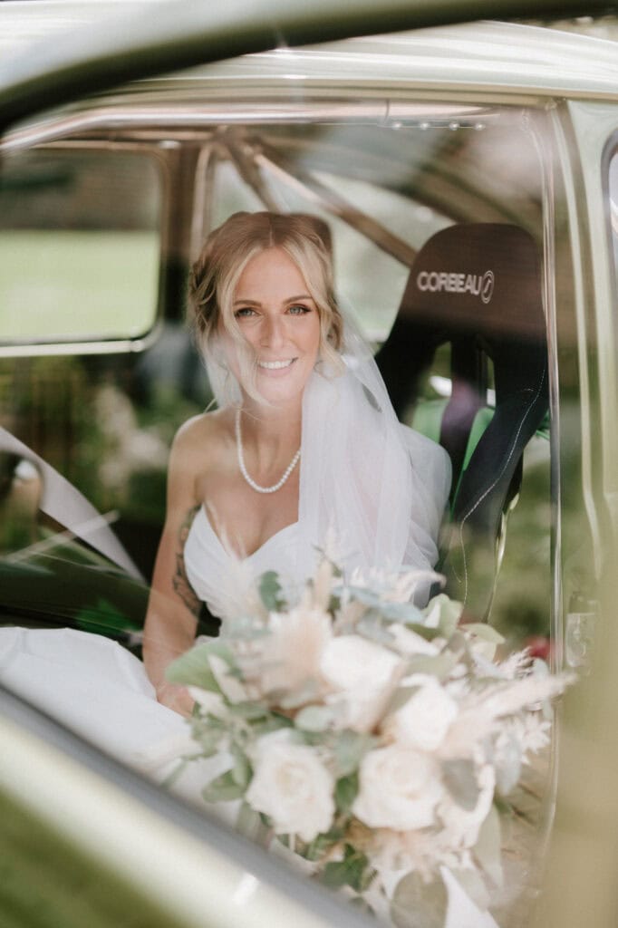A bride with blonde hair and a veil sits inside a car at The Old Kent Barn, holding a bouquet of flowers with white roses and greenery. She is smiling and wearing a white strapless wedding dress and a pearl necklace. The car seat behind her is labeled "Corbeau. Image by Pearce Wedding Photography.