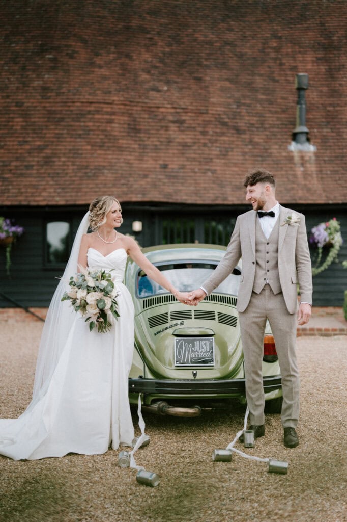 Bride and groom holding hands, standing in front of a decorated vintage car with a "Just Married" sign. The bride holds a bouquet, and both are dressed in wedding attire.