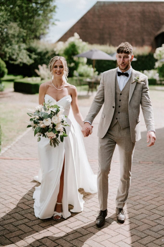 A bride in a white dress and a groom in a grey suit walk hand-in-hand outdoors on a sunny day. The bride carries a bouquet of white and green flowers.