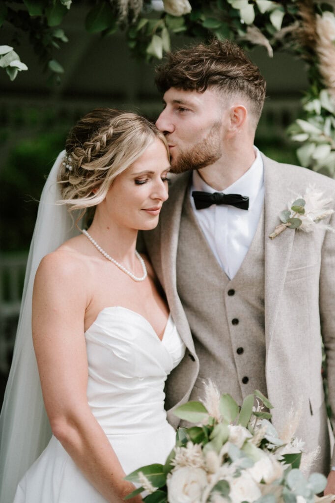 A bride and groom pose under an archway, with the groom kissing the bride's forehead. The bride wears a white gown and pearl necklace, and the groom is in a beige suit with a black bow tie.