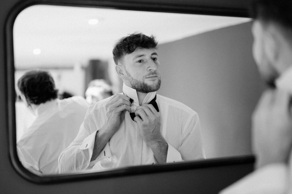 A man in a white dress shirt adjusts his bow tie while looking in a mirror. The reflection shows additional people in similar attire in the background.
