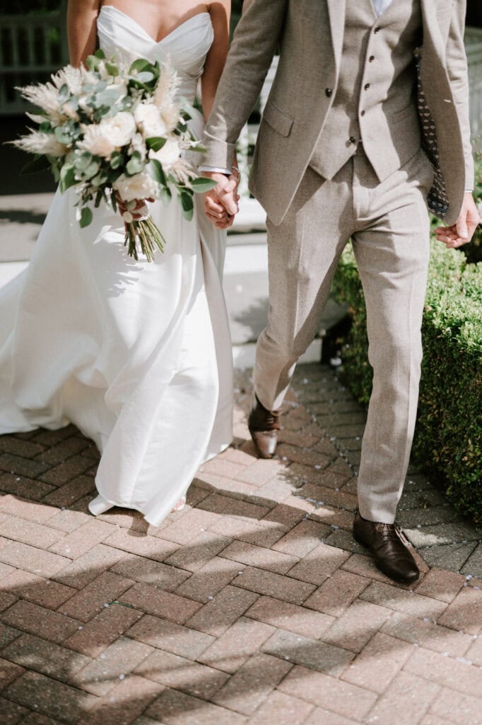 A bride in a white dress and a groom in a light grey suit walk hand in hand, with the bride holding a bouquet.