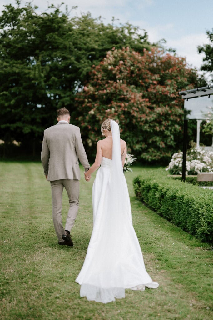 A bride in a white gown and veil, and a groom in a light grey suit, walk hand-in-hand on a grassy path surrounded by greenery and flowers.