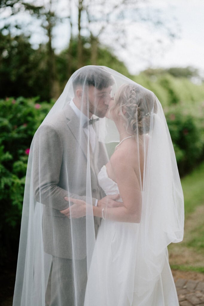 A bride and groom share a kiss outdoors, both under the bride's veil. The groom is in a gray suit and the bride is in a white strapless wedding dress. Green foliage is visible in the background.