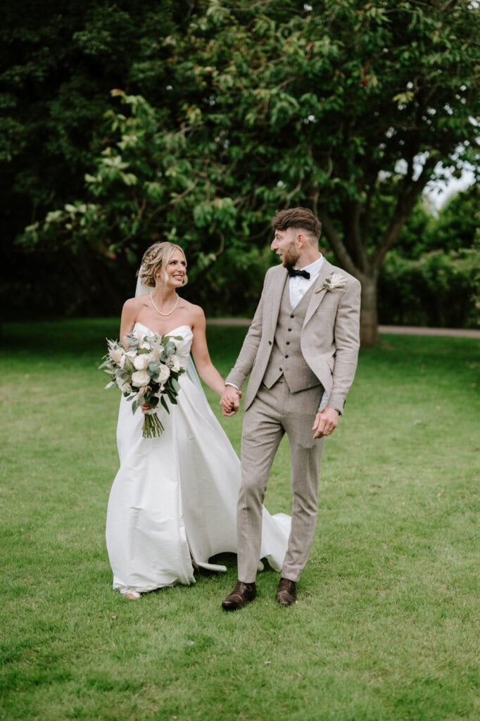 A bride in a white gown and groom in a light brown suit walk hand in hand on a grassy lawn, surrounded by trees. Both are smiling at each other. The bride holds a bouquet.