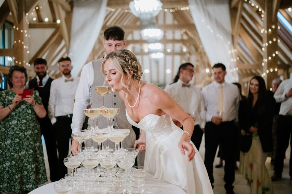 A bride in a white dress blows on a Champagne tower at a wedding reception, surrounded by guests dressed in formal attire.