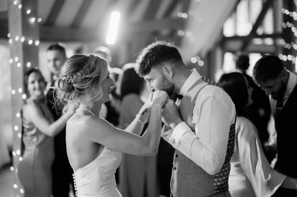 A bride and groom dance closely, with the groom kissing the bride's hand. Other guests are dancing in the background. The setting appears to be an indoor wedding reception with string lights.