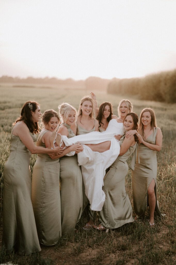 A group of seven bridesmaids in matching sage green dresses cheerfully hold up a smiling bride wrapped in a white wedding dress. They are standing outdoors in the grassy field at The Old Kent Barn during sunset, creating a warm and joyful atmosphere for the wedding. Image by Pearce Wedding Photography.