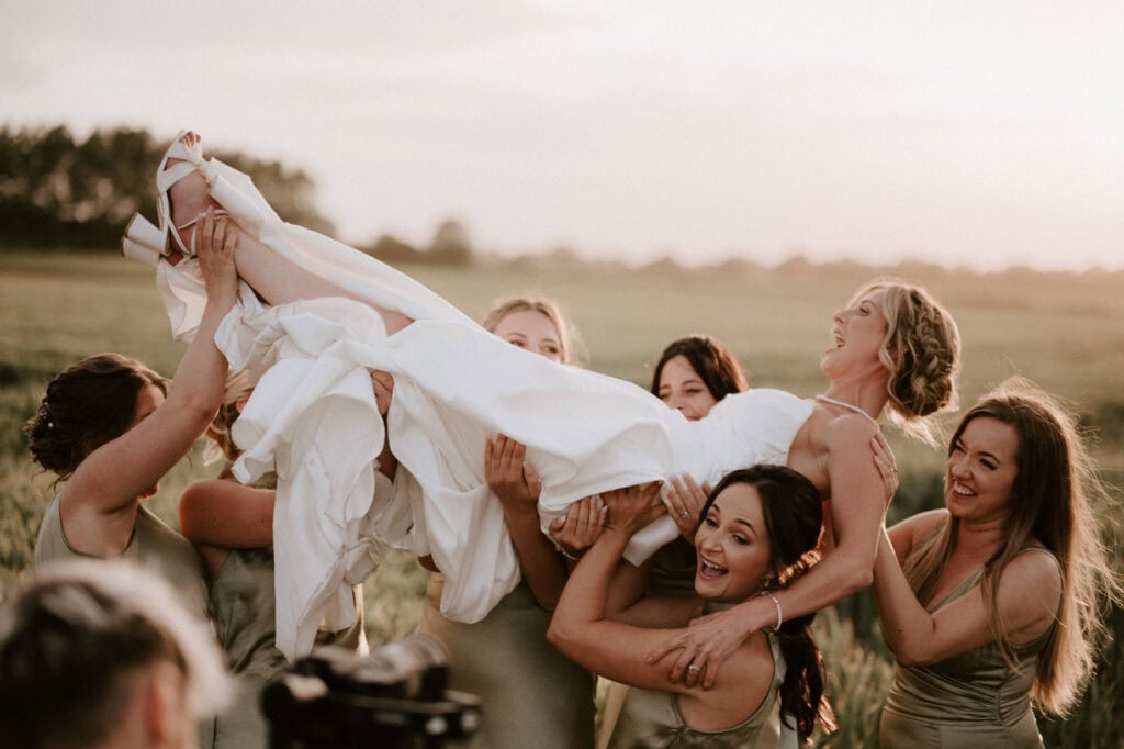 A group of bridesmaids dressed in green lifts a bride in a white gown, all smiling and laughing in a field at sunset during a beautiful wedding at The Old Kent Barn.