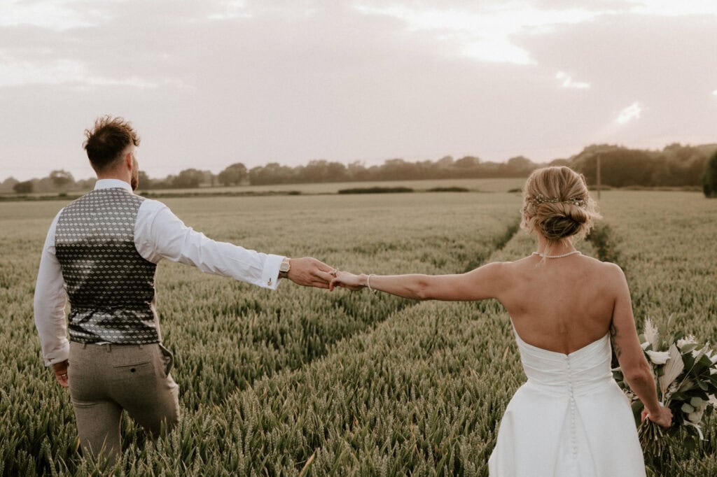 A couple in wedding attire walks hand in hand through a field of tall grass, with the sun setting in the background.