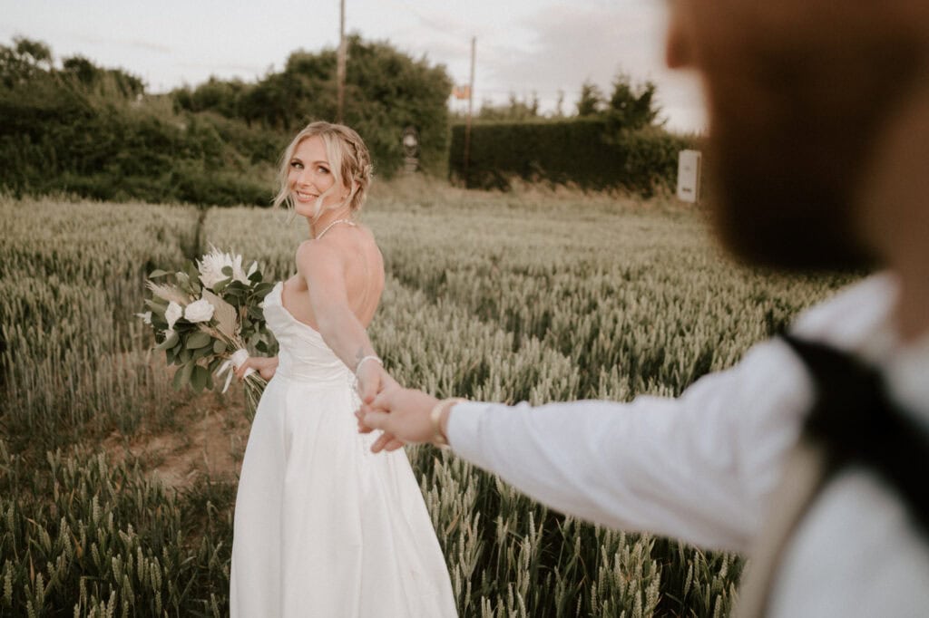 A bride in a white dress holds a bouquet and smiles while walking in a field, holding hands with a person in the foreground.
