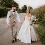 A bride and groom are joyfully walking down a rural road. The bride, in a white strapless wedding dress from Kent Bridal Shop, holds a bouquet of flowers. The groom is dressed in a light grey suit with a bow tie. Holding hands, they smile at each other, surrounded by lush green bushes and trees under a light sky. Image by Pearce Wedding Photography.