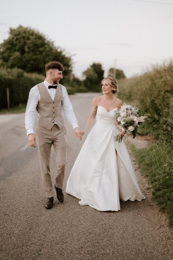 A bride and groom walk hand in hand on a road lined with greenery. The groom is in a beige suit, and the bride is in a white wedding gown holding a bouquet.
