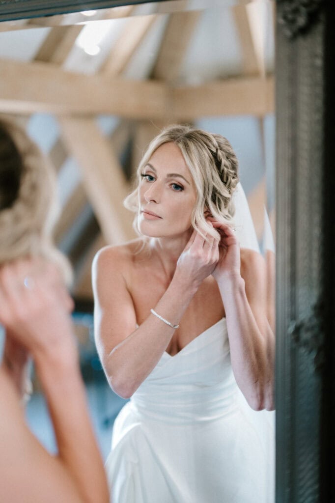 A bride with blonde hair styled in intricate braids is adjusting her earring while looking into a mirror. She is wearing a strapless white wedding dress and a thin bracelet. The softly lit room, adorned with wooden beams, perfectly captures the charm of The Old Kent Barn on her wedding day. Image by Pearce Wedding Photography.