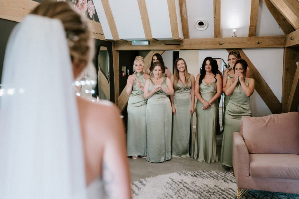 A bride in a wedding dress faces a group of six bridesmaids in sage green dresses, who appear emotional and excited in a room with wooden beams and soft lighting.