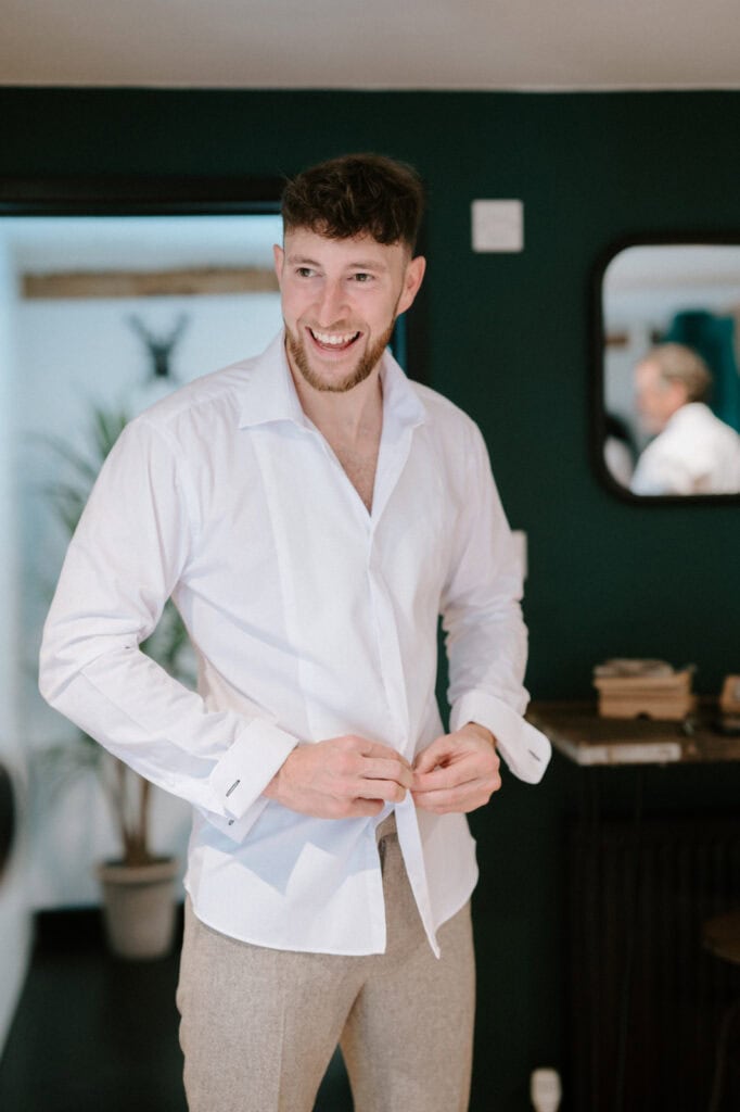 A man with a beard smiles while buttoning a white dress shirt in a room with dark green walls and a mirror in the background at The Old Kent Barn, preparing for the wedding.