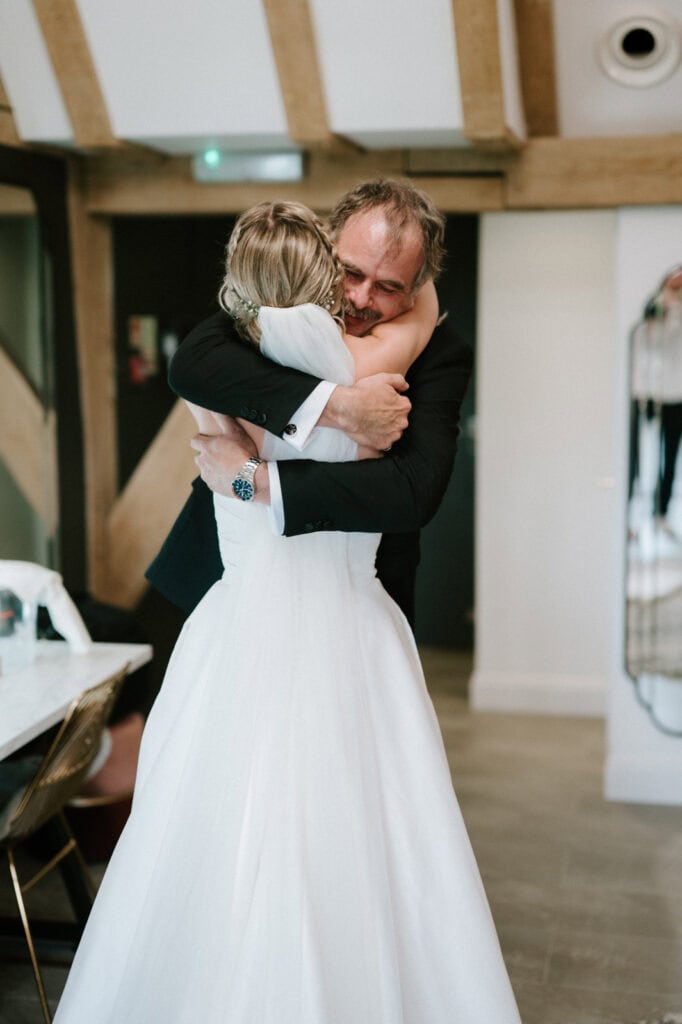 A bride in a white dress hugs a man in a suit inside a room with wooden beams and a mirror.