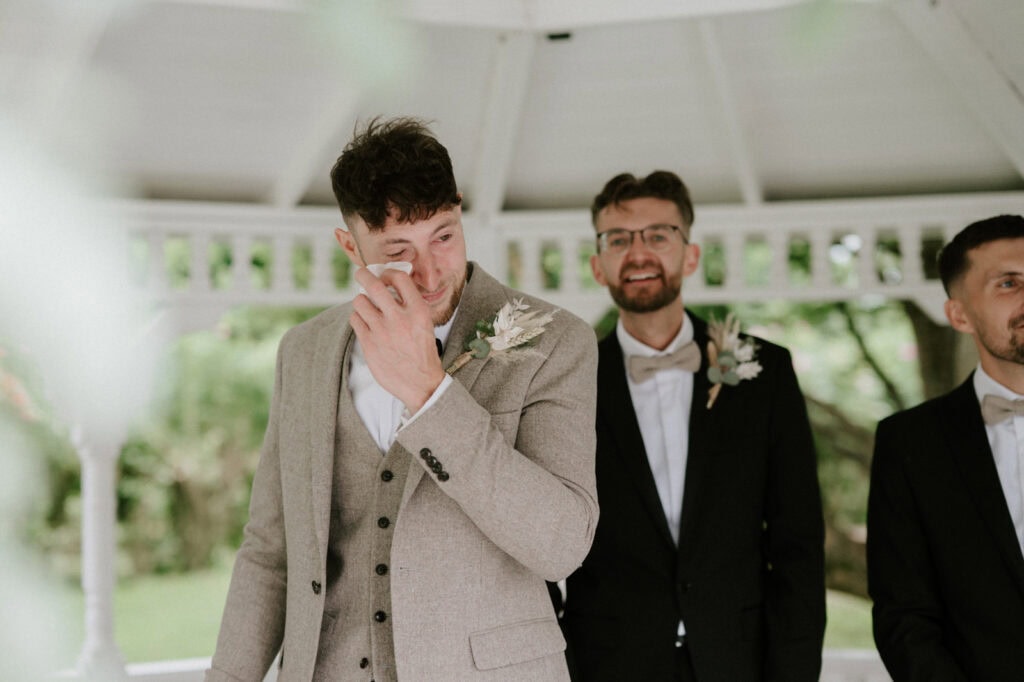 A groom in a light brown suit wipes away tears under the white gazebo at The Old Kent Barn, flanked by two groomsmen dressed in black suits and bow ties. They all wear boutonnières, with lush greenery in the background, adding to the enchanting wedding scene. Image by Pearce Wedding Photography.
