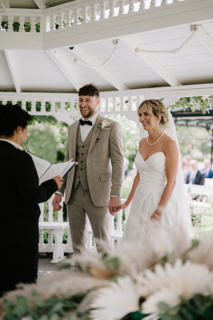 A couple stands under a gazebo at The Old Kent Barn, holding hands and facing an officiant who is reading from a book during their wedding ceremony. The groom wears a light-colored suit, and the bride wears a white dress.
