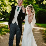 A couple stands outdoors on a sunny path, dressed in formal wedding attire. The bride smiles and raises her left hand to show her ring, while the groom smiles beside her.