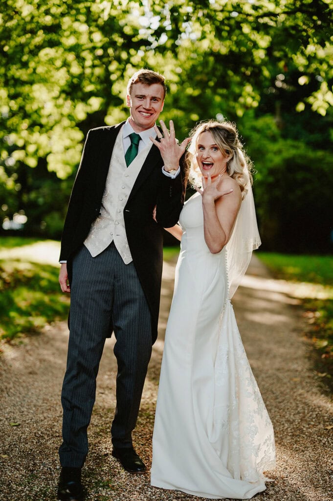 A bride and groom are outdoors, smiling and showing their wedding rings. The bride is wearing a white dress and veil, and the groom is in a suit with a green tie. They stand on a path with greenery in the background.