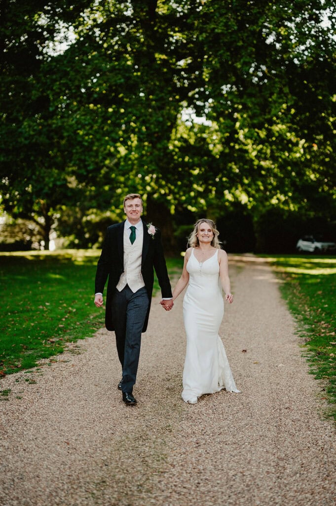 A couple, dressed in formal wedding attire, walks hand-in-hand down a tree-lined gravel path. The man is in a black suit, and the woman is in a white gown.