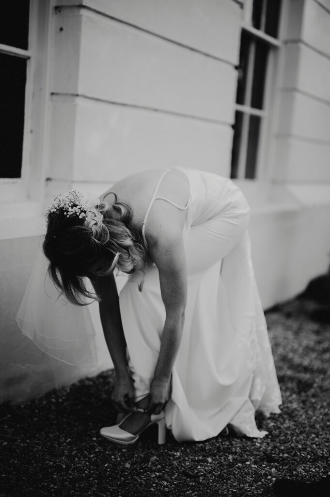 A woman in a white dress, possibly a bride, bends over to adjust her shoe. She is wearing a veil and stands outside near a building with large windows.