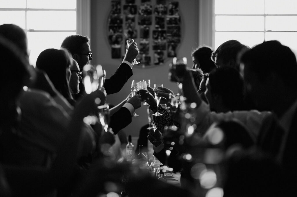 A group of people raise glasses for a toast in a dimly lit room, with a collage of photos on the wall and light coming through two windows in the background.