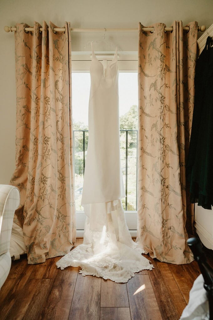 A sleeveless white wedding dress with a lace train hangs on a rod between pink floral curtains in front of a window, displaying an outdoor scene.